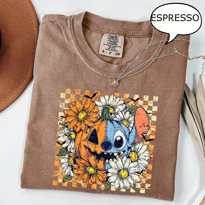 May include: Espresso-colored t-shirt with a graphic featuring a pumpkin and Stitch character surrounded by orange and white flowers. The shirt has a crew neck and short sleeves. A silver necklace is draped across the shirt.