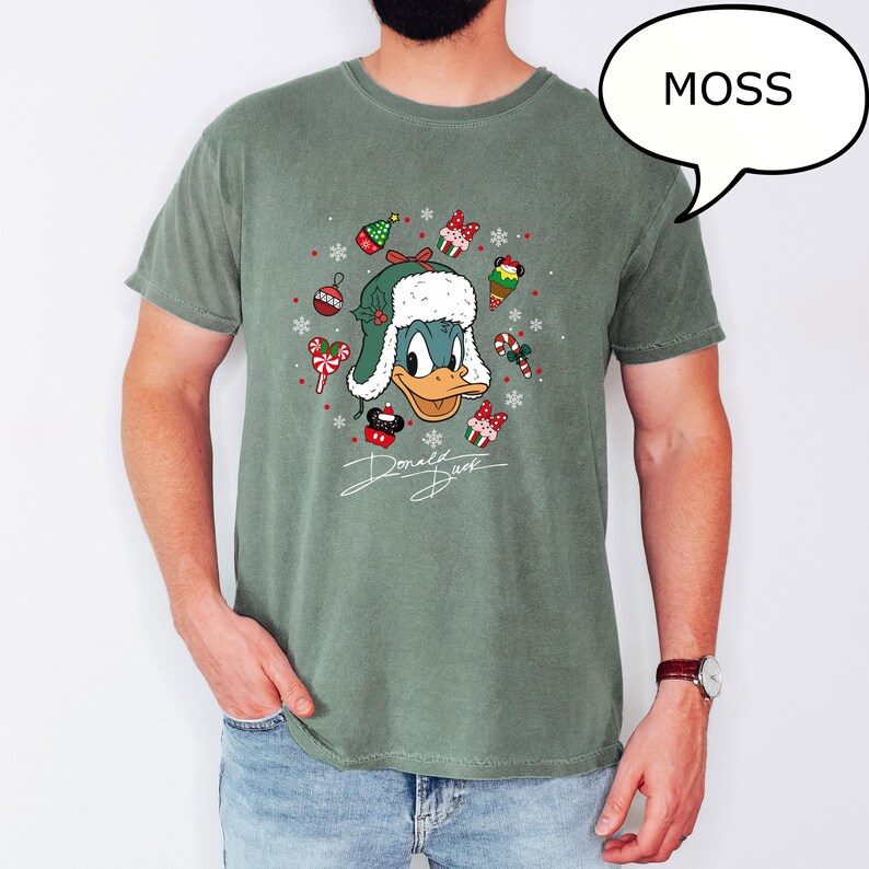 May include: A moss green t-shirt featuring a graphic of Donald Duck wearing a Christmas hat, surrounded by festive ornaments and candy canes. The text "Donald Duck" is written in a cursive font below the graphic. A speech bubble in the upper right corner says "MOSS".