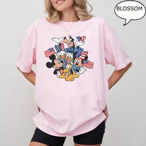 May include: A light pink t-shirt featuring a graphic of Mickey Mouse, Donald Duck, Goofy, Pluto, and Minnie Mouse, each holding an American flag. The word "BLOSSOM" is in a speech bubble.