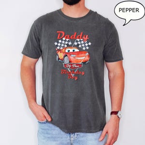 Puede incluir: Una camiseta gris oscuro con un coche de carreras rojo de dibujos animados y el texto "Daddy Of The Birthday Boy" impreso en ella.