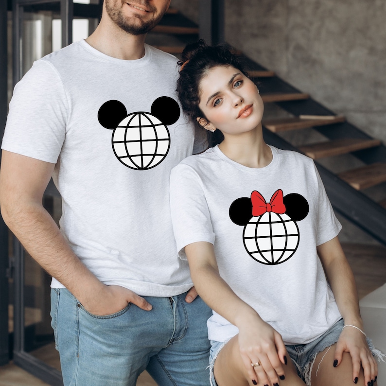 K&ouml;nnte beinhalten: Ein Paar tr&auml;gt wei&szlig;e T-Shirts mit einer schwarzen und wei&szlig;en Globusgrafik. Das Shirt der Frau hat eine rote Schleife auf dem Globus. Die Globusgrafik ist ein stilisierter Mickey-Maus-Kopf.