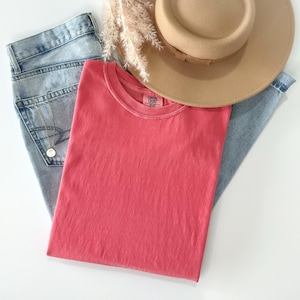 May include: A coral pink crew neck t-shirt with a Comfort Colors label, light blue denim jeans, a tan wide-brimmed hat, a gold chain necklace with a circular pendant, and a sprig of dried white pampas grass arranged on a plain white surface.