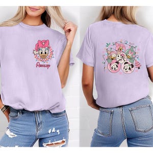K&ouml;nnte beinhalten: Lilafarbenes T-Shirt mit Daisy Duck mit rosa Schleife und Blumenkranz, darunter der Name "Ashley". Auf der R&uuml;ckseite sind Disney-Figuren in Weihnachtsbaumkugel-Designs zu sehen, darunter Mickey und Minnie Mouse.