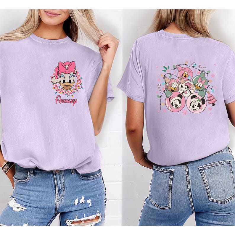 K&ouml;nnte beinhalten: Lilafarbenes T-Shirt mit Daisy Duck mit rosa Schleife und Blumenkranz, darunter der Name "Ashley". Auf der R&uuml;ckseite sind Disney-Figuren in Weihnachtsbaumkugel-Designs zu sehen, darunter Mickey und Minnie Mouse.