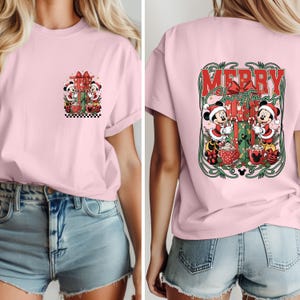 May include: Pale pink t-shirt with a Christmas design featuring Mickey and Minnie Mouse. The front has a gift box and festive details. The back of the shirt displays "MERRY CHRISTMAS" in a decorative frame with the characters.