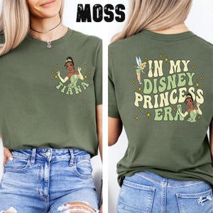 May include: A green t-shirt with a graphic of Tiana from Disney's Princess and the Frog on the front and the text "In My Disney Princess Era" on the back.