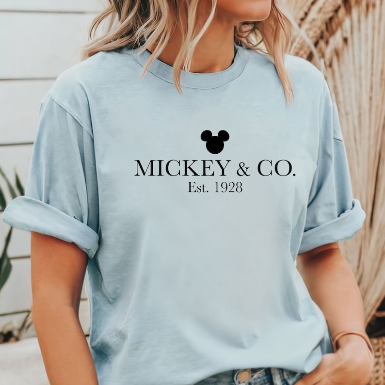 Puede incluir: Una camiseta azul claro con una silueta negra de Mickey Mouse y el texto "MICKEY & CO. Est. 1928"
