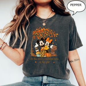 May include: Dark gray t-shirt featuring Mickey and Minnie Mouse under a tree with fall foliage. The graphic includes pumpkins and the text "It's the most wonderful time of the year". A speech bubble says "PEPPER".