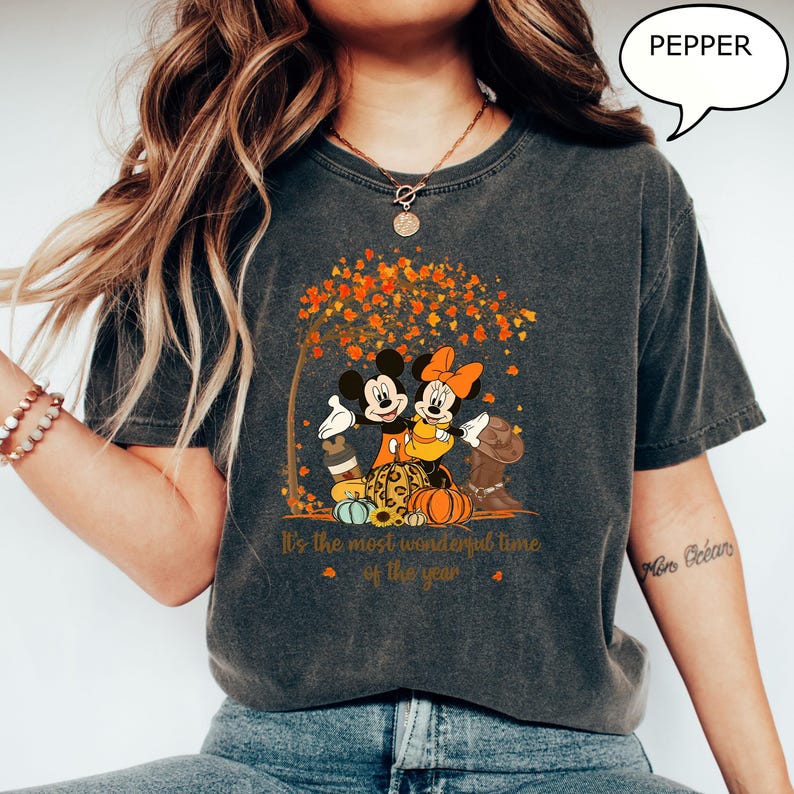 May include: Dark gray t-shirt featuring Mickey and Minnie Mouse under a tree with fall foliage. The graphic includes pumpkins and the text "It's the most wonderful time of the year". A speech bubble says "PEPPER".