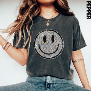 May include: A gray t-shirt with a distressed, leopard print smiley face graphic. The shirt has a relaxed fit and a crew neck.