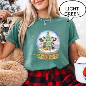 May include: Light green t-shirt featuring a snow globe design with Mickey and Minnie Mouse in Christmas attire. The snow globe includes a Christmas tree, gifts, and candy. The text "LIGHT GREEN" is in a speech bubble.