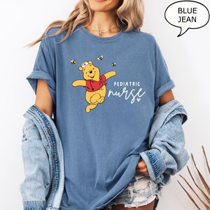 May include: Blue jean t-shirt featuring a cartoon bear wearing a nurse's hat and red vest, with bees. The text reads "Pediatric Nurse" in white script. The shirt has rolled-up sleeves.