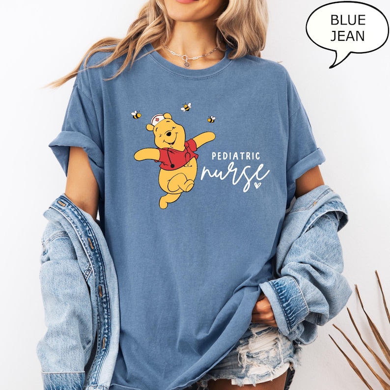May include: Blue jean t-shirt featuring a cartoon bear wearing a nurse's hat and red vest, with bees. The text reads "Pediatric Nurse" in white script. The shirt has rolled-up sleeves.
