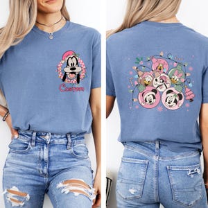 K&ouml;nnte beinhalten: Blaues T-Shirt mit einem Cartoon-Bild von Goofy mit Weihnachtsm&uuml;tze und dem Wort "Custom". Auf der R&uuml;ckseite des Shirts sind Disney-Figuren in Weihnachtskugeln abgebildet. Das Shirt besteht aus weichem Material.