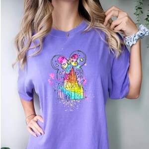 Puede incluir: Una camiseta morada con un boceto arco&iacute;ris del castillo de Cenicienta con orejas de Mickey Mouse en la parte superior. El castillo est&aacute; rodeado de salpicaduras de pintura de colores.