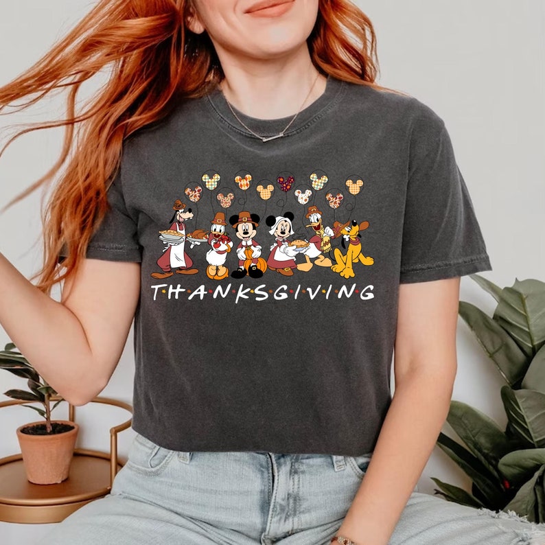 May include: A dark gray t-shirt with a Thanksgiving theme featuring Mickey Mouse and friends. The shirt has a line of Mickey Mouse heads with balloons above a line of characters including Mickey Mouse, Minnie Mouse, Donald Duck, Goofy, and Pluto. The text "THANKSGIVING" is below the characters.