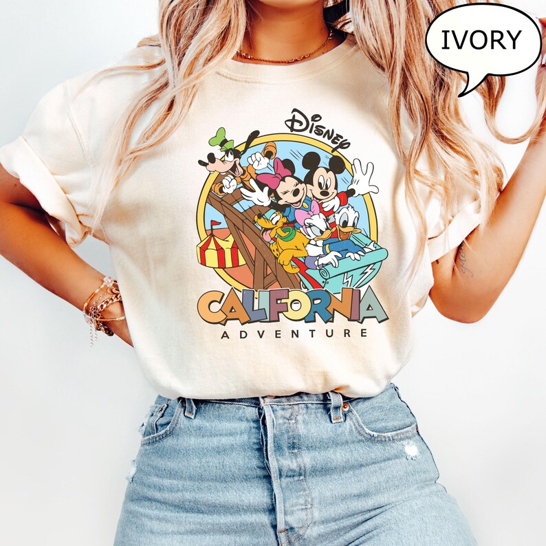 Puede incluir: Una camiseta blanca con un gr&aacute;fico colorido de Mickey Mouse y sus amigos montando una monta&ntilde;a rusa. El gr&aacute;fico incluye el texto "Disney California Adventure".