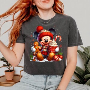 Puede incluir: Una camiseta negra recortada con un gr&aacute;fico navide&ntilde;o que presenta a Mickey Mouse con un gorro de Pap&aacute; Noel rojo y blanco, sosteniendo un hombre de jengibre y una taza de chocolate caliente con crema batida.