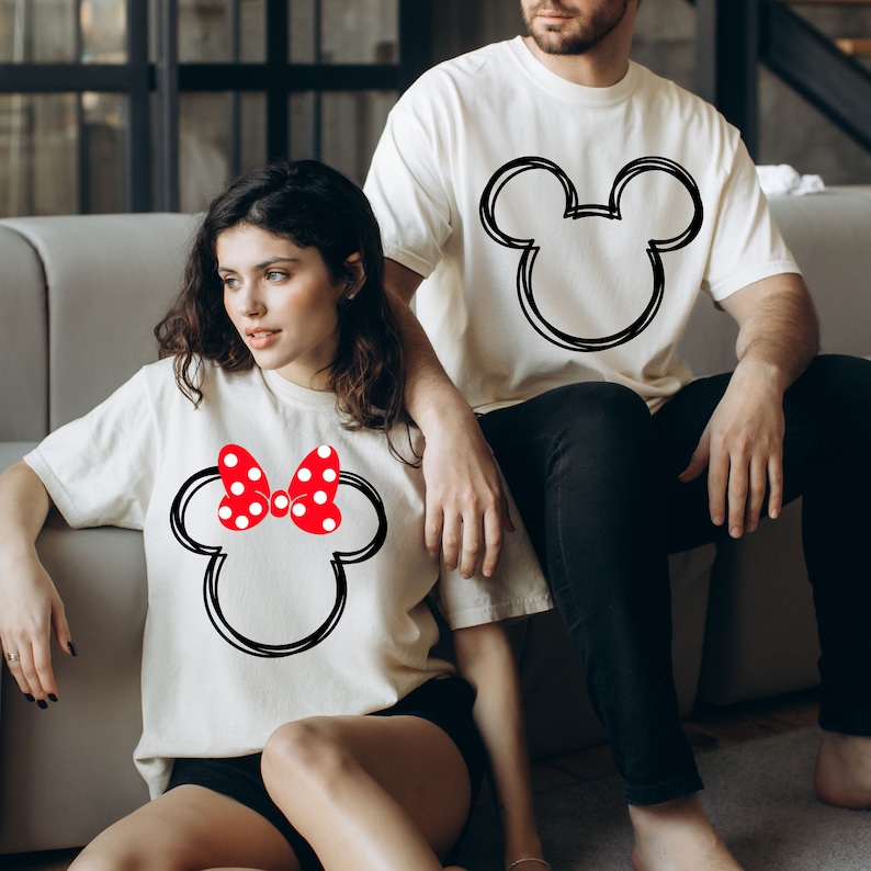 May include: A couple wearing white t-shirts with a black and white Mickey Mouse and Minnie Mouse design. The Minnie Mouse shirt has a red bow with white polka dots.