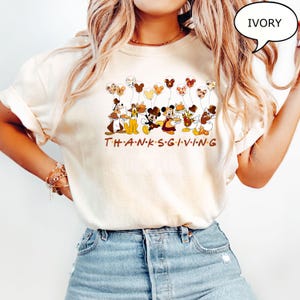 May include: Ivory-colored t-shirt featuring Disney characters holding balloons and Thanksgiving food. The word "THANKSGIVING" is printed in brown letters below the characters. The shirt has a relaxed fit.