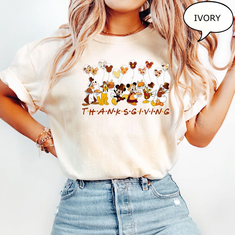 May include: Ivory-colored t-shirt featuring Disney characters holding balloons and Thanksgiving food. The word "THANKSGIVING" is printed in brown letters below the characters. The shirt has a relaxed fit.