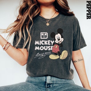 May include: A dark gray t-shirt with a vintage Mickey Mouse graphic. The graphic features Mickey Mouse in his signature red shorts and yellow shoes. The text "Mickey Mouse" is printed in white below the graphic. The shirt has a crew neck and short sleeves.