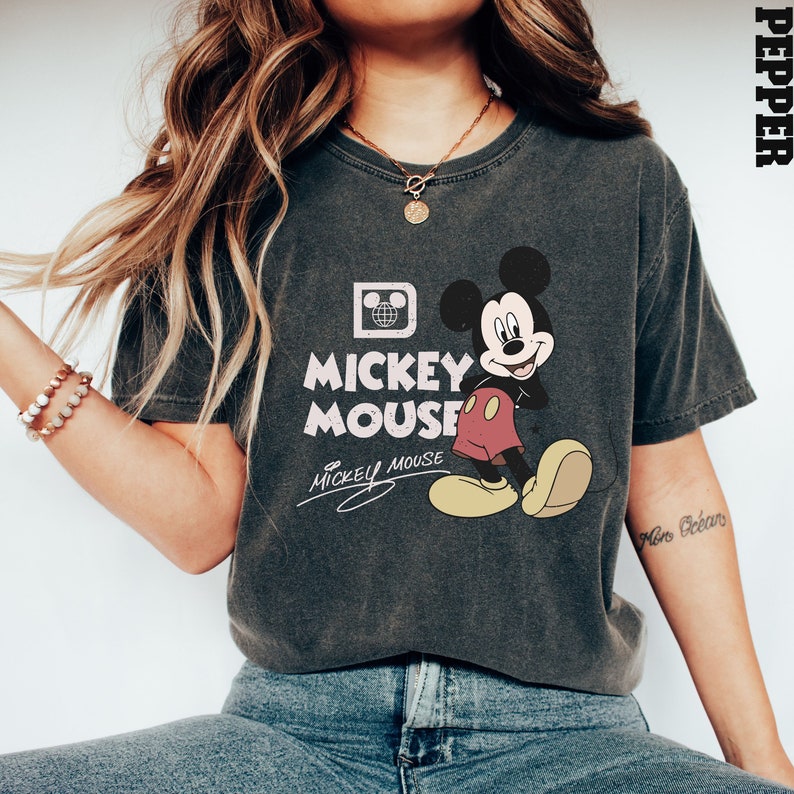 May include: A dark gray t-shirt with a vintage Mickey Mouse graphic. The graphic features Mickey Mouse in his signature red shorts and yellow shoes. The text "Mickey Mouse" is printed in white below the graphic. The shirt has a crew neck and short sleeves.