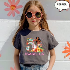 May include: A gray t-shirt featuring a colorful graphic of a cartoon character, the number 5, and the name "ISABELLA" in pink. The girl in the image is wearing pink sunglasses and is standing in front of a white wall with flower decorations.