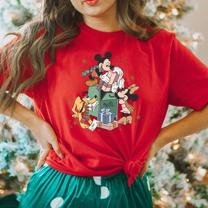 May include: Red t-shirt featuring a Christmas-themed graphic of Mickey Mouse, Minnie Mouse, and Pluto delivering presents to a mailbox. The graphic is in a vintage style with gold stars and a green mailbox.