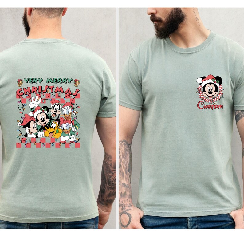 May include: Sage green t-shirt featuring a Christmas-themed graphic. The front displays a Mickey Mouse design with a Santa hat and the word "Custom". The back has a larger graphic with Mickey, Minnie, Donald, Goofy, and Pluto with the text "Very Merry Christmas".