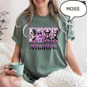 May include: A moss green t-shirt featuring a graphic of four cartoon villains with pink accents. The text "Sweetie I'm OK with being the Villain" is below the graphic. A speech bubble with the word "MOSS" is in the upper right.