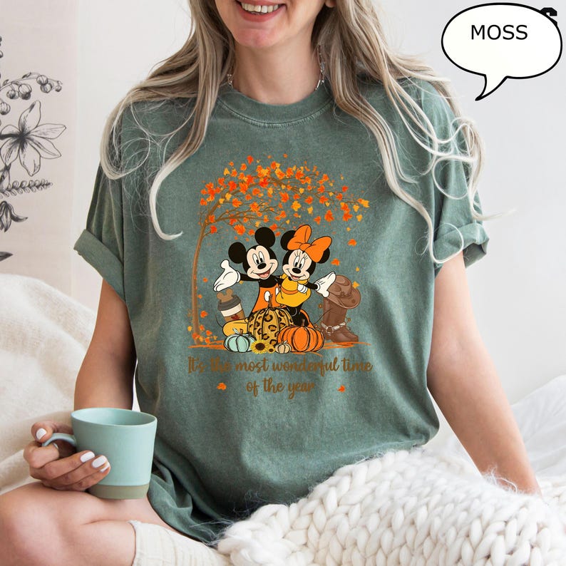 May include: Moss green t-shirt featuring Mickey and Minnie Mouse embracing under a tree with autumn leaves. The graphic includes pumpkins and the text "It's the most wonderful time of the year."