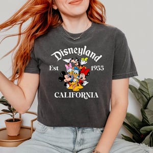 Puede incluir: Una camiseta gris oscuro recortada con un estampado gr&aacute;fico blanco de Mickey Mouse y sus amigos. El gr&aacute;fico incluye el texto "Disneyland Est 1955 CALIFORNIA".