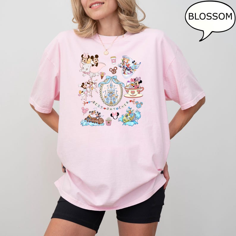 Puede incluir: Camiseta rosa claro con un dise&ntilde;o gr&aacute;fico colorido con personajes de dibujos animados y el texto "BEST DAY EVER". El dise&ntilde;o incluye im&aacute;genes de un castillo, tazas de t&eacute; y otros elementos caprichosos. La camiseta es de corte holgado.