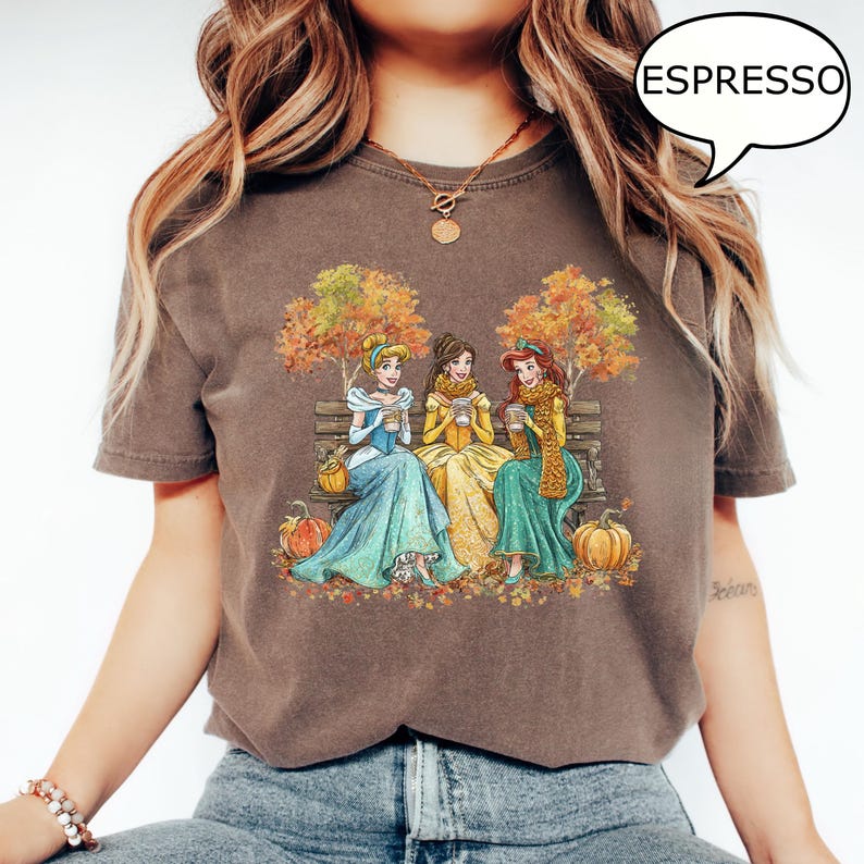 May include: Espresso-colored t-shirt featuring a graphic of three princesses sitting on a bench, enjoying coffee in the fall. The princesses are wearing colorful gowns and scarves, with pumpkins and autumn leaves scattered around them.