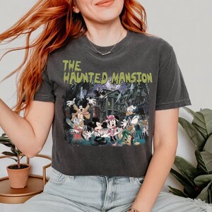 May include: A black cropped t-shirt with a vintage-style graphic of Mickey Mouse and friends in front of the Haunted Mansion. The text "THE HAUNTED MANSION" is printed in green on the shirt.