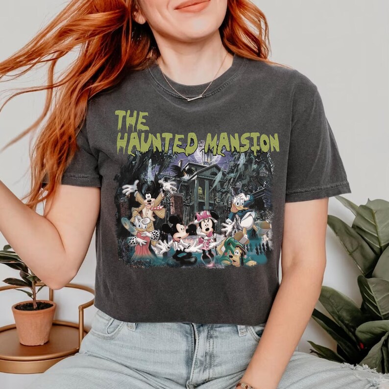 May include: A black cropped t-shirt with a vintage-style graphic of Mickey Mouse and friends in front of the Haunted Mansion. The text "THE HAUNTED MANSION" is printed in green on the shirt.
