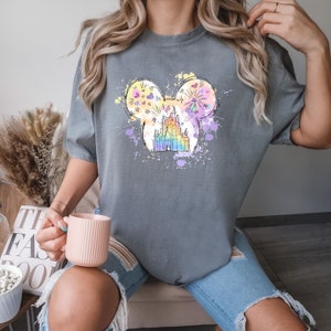 Puede incluir: Una camiseta gris con un gr&aacute;fico colorido de un castillo en una silueta de Mickey Mouse. El castillo est&aacute; rodeado de salpicaduras de pintura coloridas y fuegos artificiales.