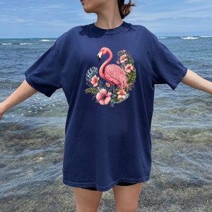K&ouml;nnte beinhalten: Ein marineblaues T-Shirt mit einem pinkfarbenen Flamingo und einem Blumendesign. Der Flamingo steht auf einem Bein und hat den Kopf zur Seite gedreht. Die Blumen sind pink und wei&szlig;.