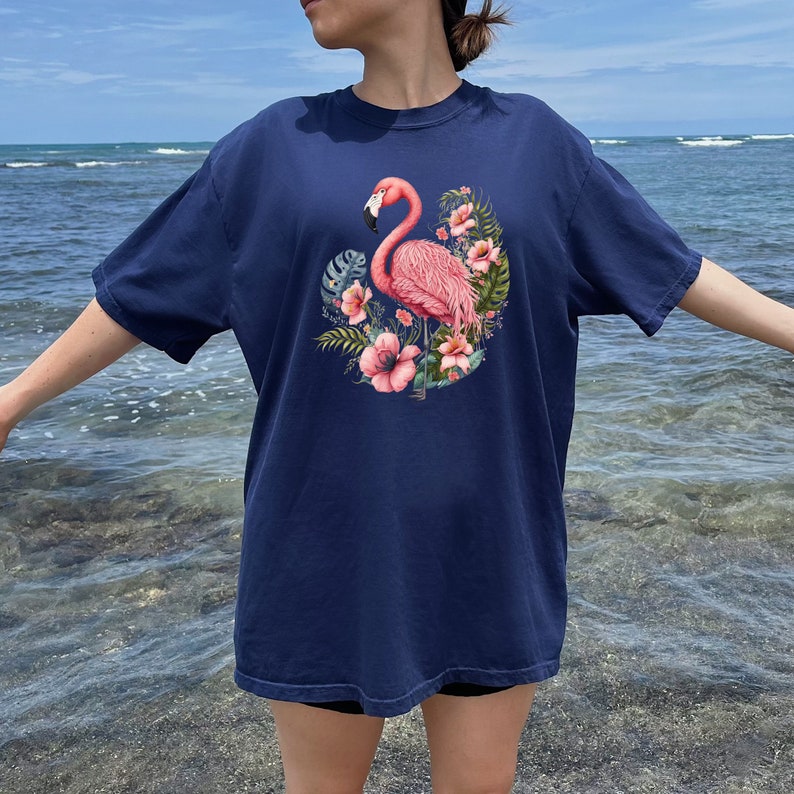 K&ouml;nnte beinhalten: Ein marineblaues T-Shirt mit einem pinkfarbenen Flamingo und einem Blumendesign. Der Flamingo steht auf einem Bein und hat den Kopf zur Seite gedreht. Die Blumen sind pink und wei&szlig;.