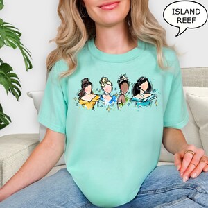 May include: A light turquoise t-shirt featuring a watercolor illustration of four princesses. The princesses are depicted with various hairstyles and gowns, accented with gold and blue stars. The text "ISLAND REEF" is in a speech bubble.