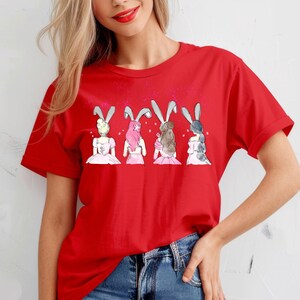 Puede incluir: Camiseta roja con un gr&aacute;fico de cuatro mujeres con orejas de conejo y vestidos rosas. Las mujeres est&aacute;n de espaldas al espectador.