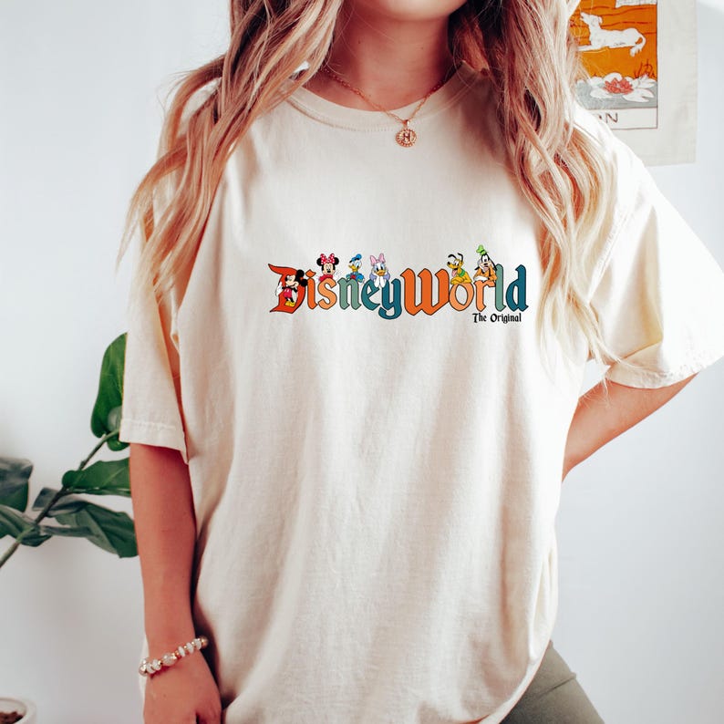 K&ouml;nnte beinhalten: Ein wei&szlig;es T-Shirt mit einem bunten Disney World Logo, auf dem Mickey Mouse, Minnie Mouse, Donald Duck, Goofy, Pluto und Chip und Dale zu sehen sind. Unter dem Logo steht der Text "The Original."