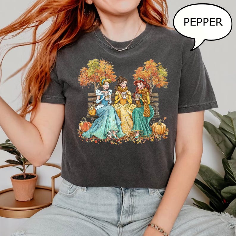 May include: Dark gray t-shirt featuring an illustration of three princesses sitting on a bench, enjoying warm beverages. The scene is set in autumn, with pumpkins and colorful leaves. The word "PEPPER" is in a speech bubble.