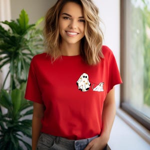 May include: A red t-shirt featuring a white ghost and a white dog wearing a ghost costume. The ghost is holding a leash attached to the dog.