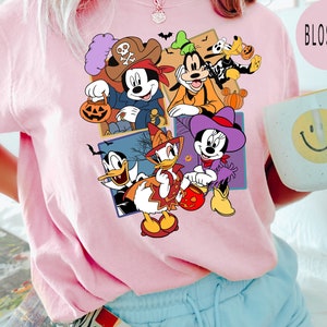 K&ouml;nnte beinhalten: Ein pinkfarbenes T-Shirt mit einem bunten Grafikdruck von Mickey Mouse, Minnie Mouse, Donald Duck, Goofy und Pluto in Halloween-Kost&uuml;men. Die Figuren halten S&uuml;&szlig;igkeiteneimer und K&uuml;rbisse.