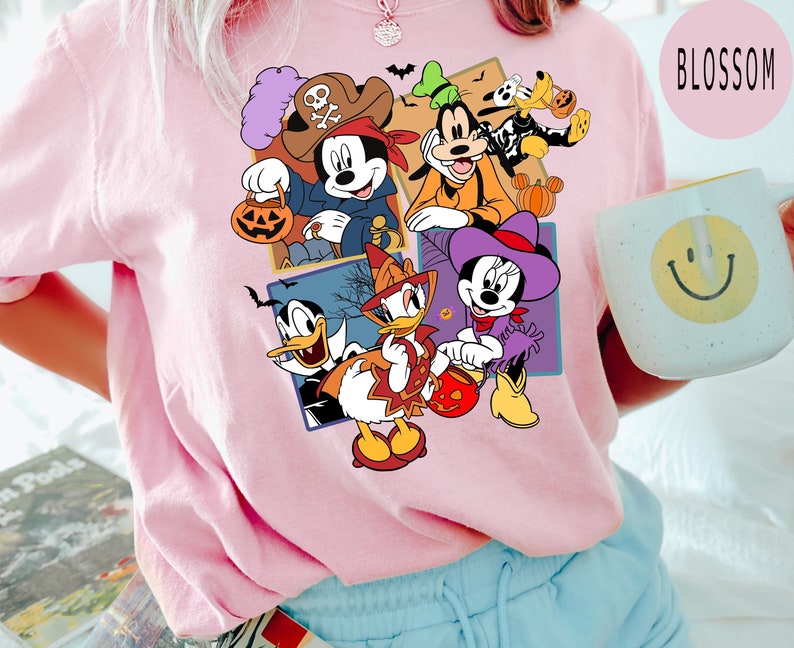 K&ouml;nnte beinhalten: Ein pinkfarbenes T-Shirt mit einem bunten Grafikdruck von Mickey Mouse, Minnie Mouse, Donald Duck, Goofy und Pluto in Halloween-Kost&uuml;men. Die Figuren halten S&uuml;&szlig;igkeiteneimer und K&uuml;rbisse.