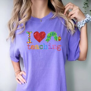 Op de afbeelding: Paars T-shirt met een kleurrijke afbeelding met de tekst "Ik hou van lesgeven" met een rood hart en een groene rups.