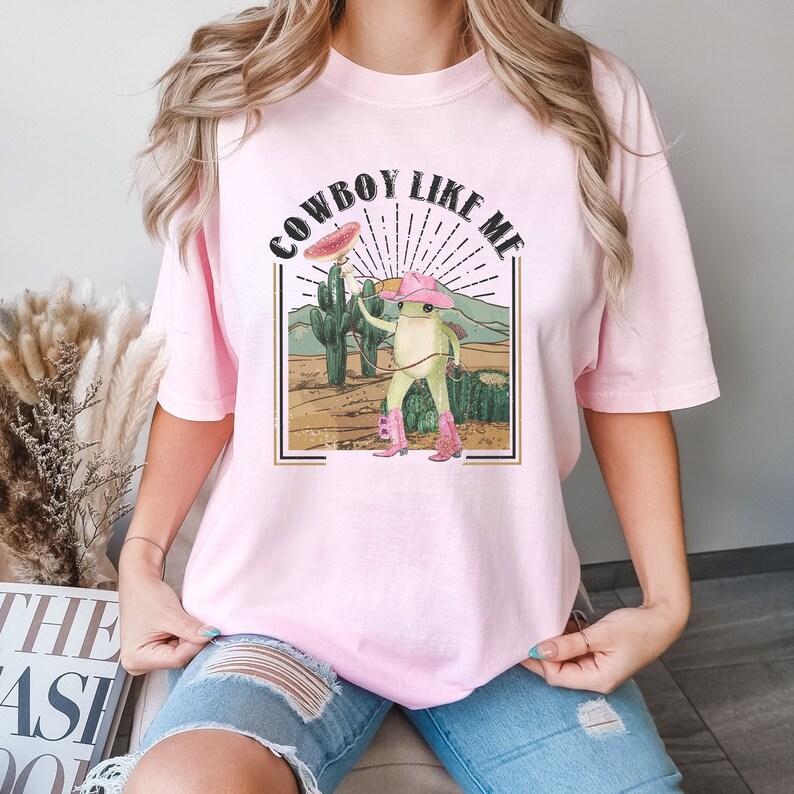 May include: A pink t-shirt with a graphic of a frog wearing a cowboy hat and boots. The text "COWBOY LIKE ME" is printed on the shirt.