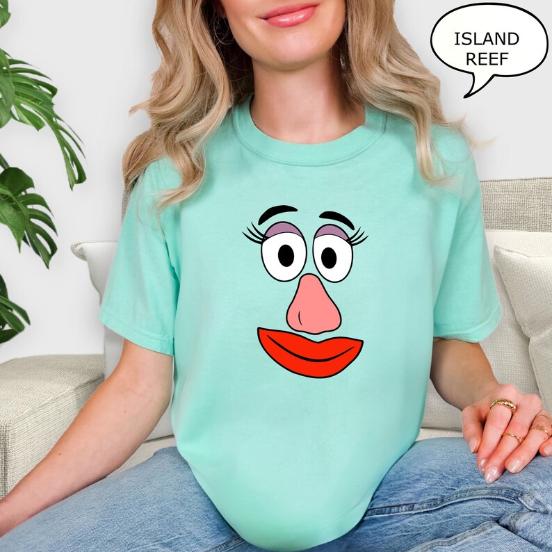 May include: A light teal t-shirt featuring a cartoon face with large eyes, a pink nose, and a red mouth. The text "ISLAND REEF" is in a speech bubble. The shirt is a casual style.
